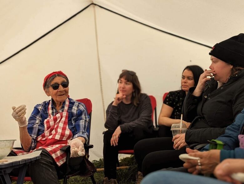 An elderly woman in a plaid shirt sits in a chair and speaks to a group of students who are leaning in and listening. 