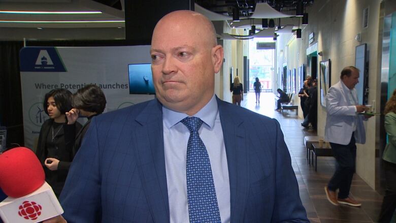 A bald man in a blue suit looking to the left side of the frame at a reporter.