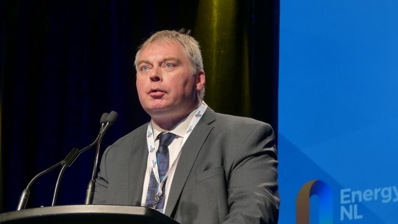 A man with grey hair, grey suit and a lanyard around his neck speaks at a microphone.