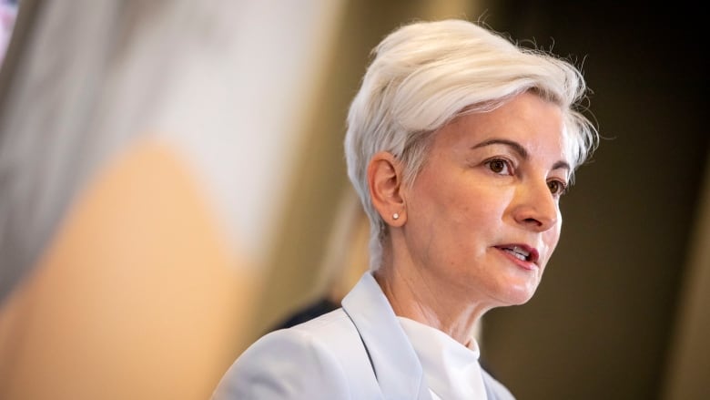 An older, handsomely attired woman with silver hair speaks in front of an amber and white backdrop