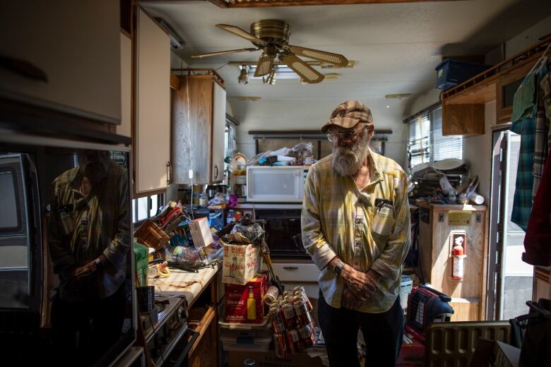 An older with a white beard wearing a baseball cap and an open-necked checked yellow and white cotton shirt looks around his trailer, which is filled with household items.