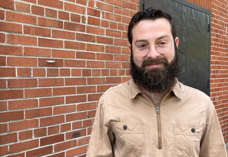 A man wearing glasses, a beard and a beige collared shirt, smiles at the camera, standing in front of a brick wall. 