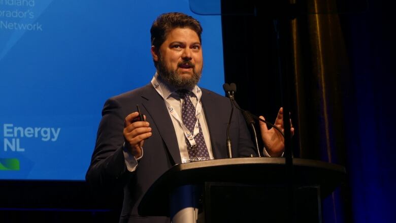 A man with a beard and black hair, wearing a suit, standing at a podium in front of a microphone.