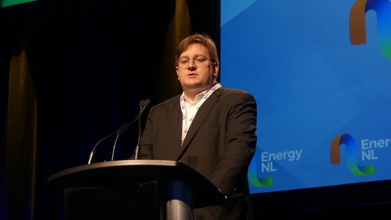 A man with thin glasses and medium length brown hair, standing at a podium and speaking into a microphone.