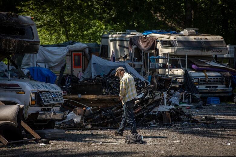 An older man in a white and yellow checked shirt walks around a parking lot filled with rundown vehicles and debris.