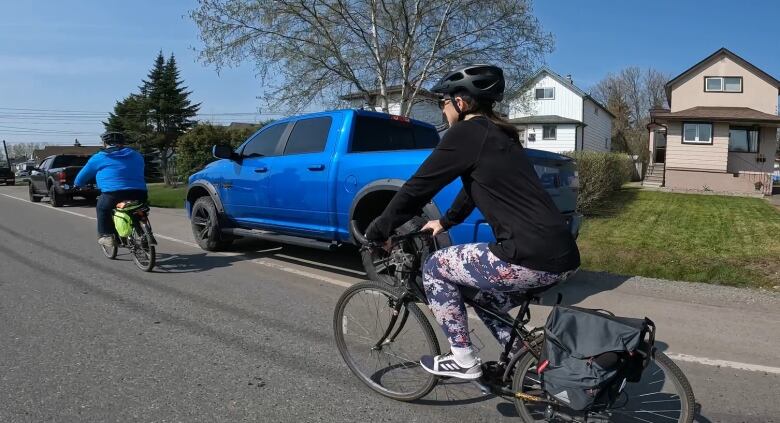 A blue truck is parked on the road shoulder while Ken Shields and CBC reporter Heather Kitching cycle around it. 