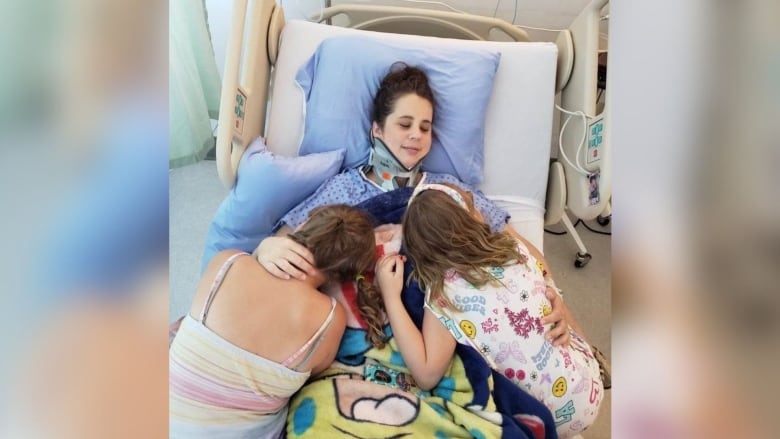 A woman in a neck brace embraces two children while on a hospital bed.