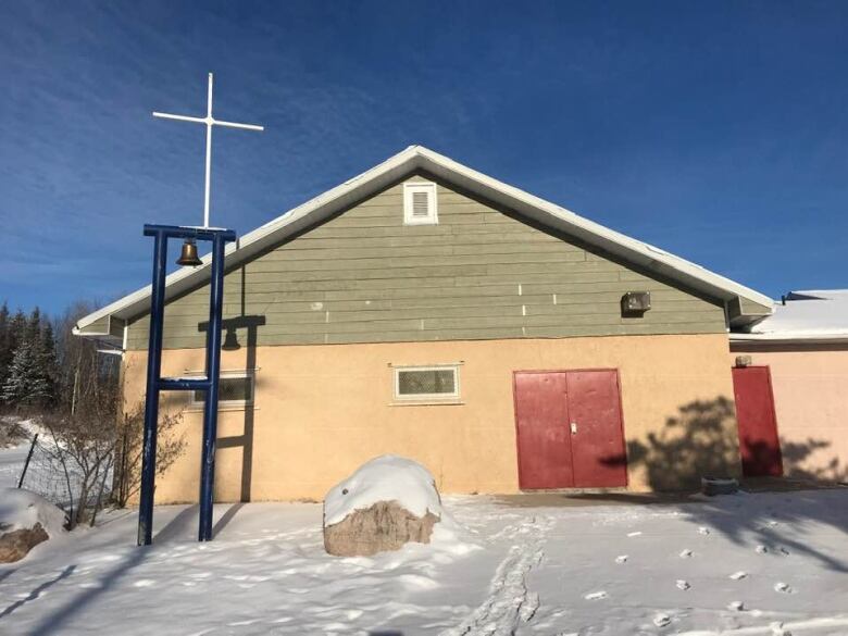 A simple building with a white cross and bell erected in front. Snow is on the ground.