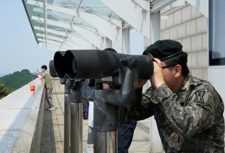 A soldier looks through large binoculars outside a building.