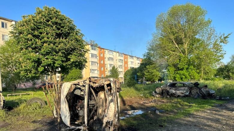 A damaged vehicle turned on its side is shown in a field, with a low-rise building in the background.