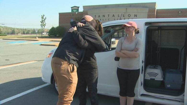 A man hugs a woman in front of a white van. Another woman watches and smiles. 