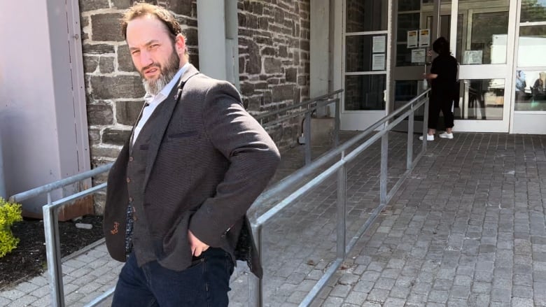 A man with thinning dark hair and a greying beard grimaces at the camera. He's wearing  a suit jacket and jeans. It's a sunny day and he's outside a glass and brick building.