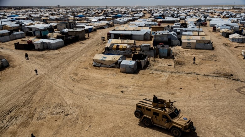 Make shift shelters dot the horizon as a military truck drives by in the foreground.