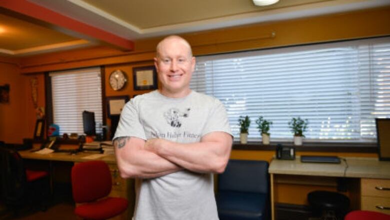 A bald man in a grey T-shirt and blue athletic shorts crosses his arms and smiles for a photo.