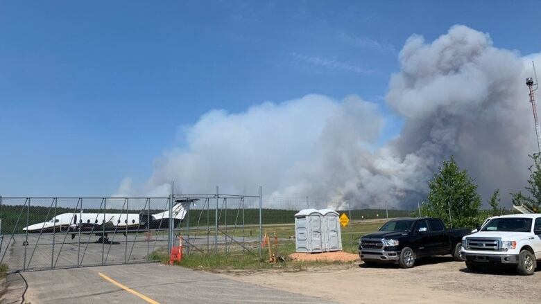 A small plane on a runway with nearby vehicles as a large plume of smoke can be seen on the horizon.