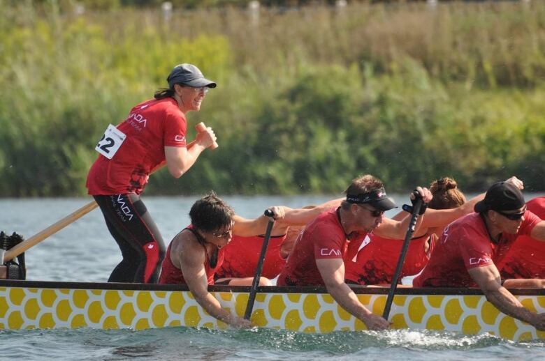 Men in Team Canada t-shirts paddle a dragon boat with determination. A woman in a ball cap stands at the back of the boat, steering and smiling. 