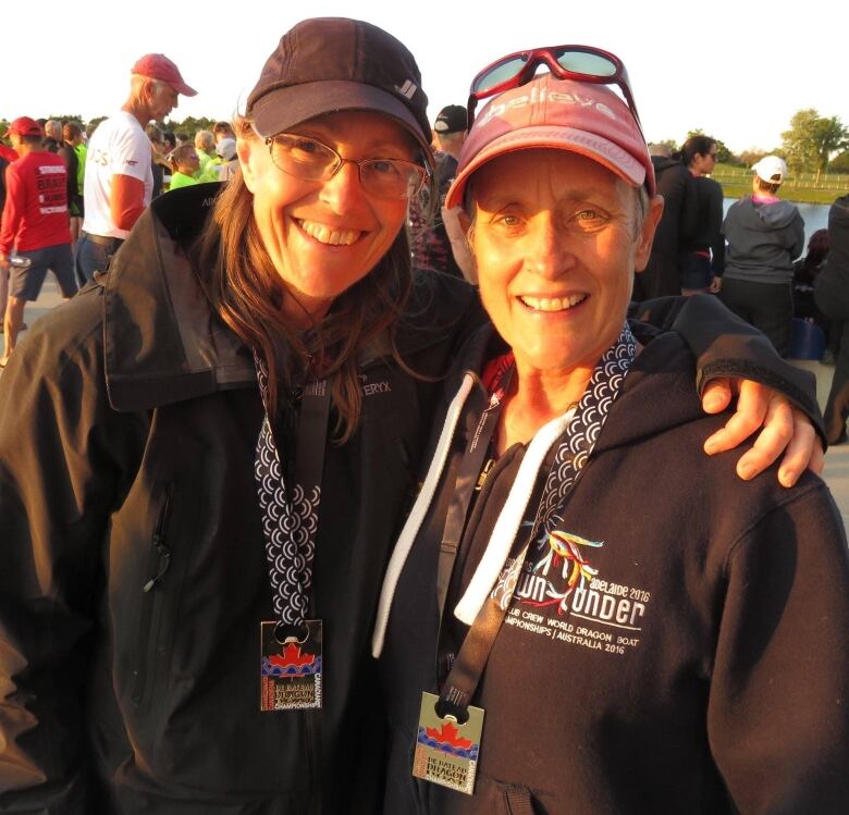 Two women stand shoulder to shoulder, wearing matching, black jackets and medals. One has their arm around the other. 