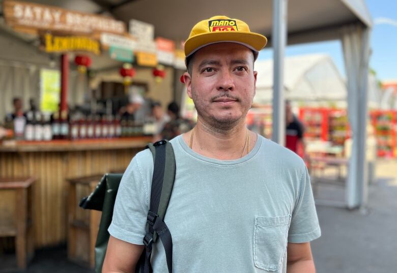 A man stands in an outdoor market wearing a yellow cap, blue shirt and backpack.