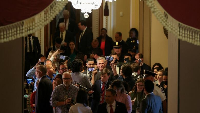 A large group of people is visible in a room. Some are using recording devices. A large chandelier is overhead.