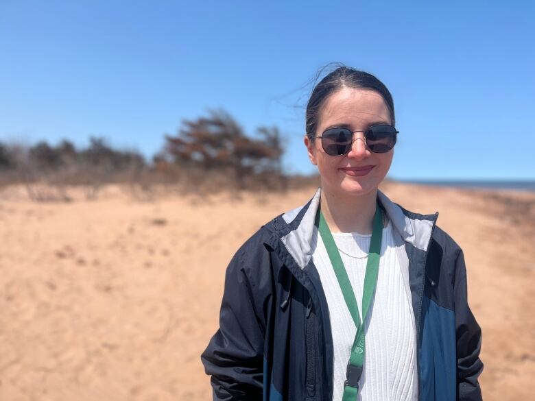 A woman stands on a beach smiling. She is wearing sunglasses, and a white top under a blue jacket. 