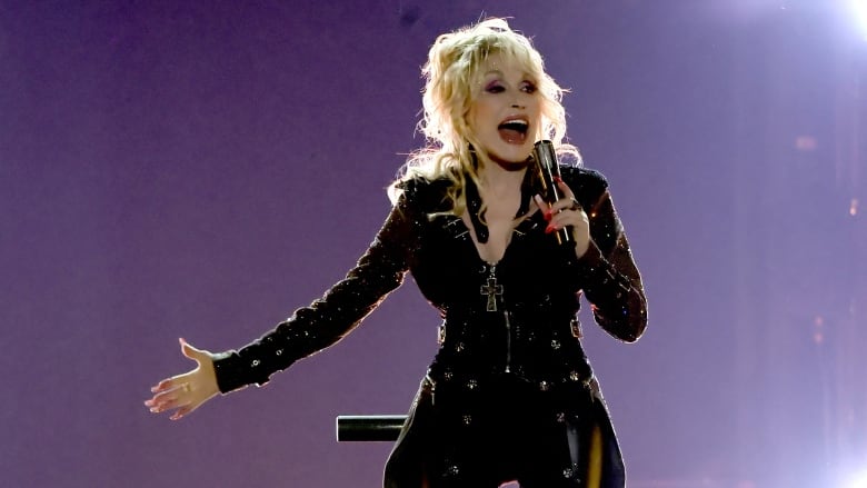 A woman with high-piled blond hair and a dark outfit performs on a stage.