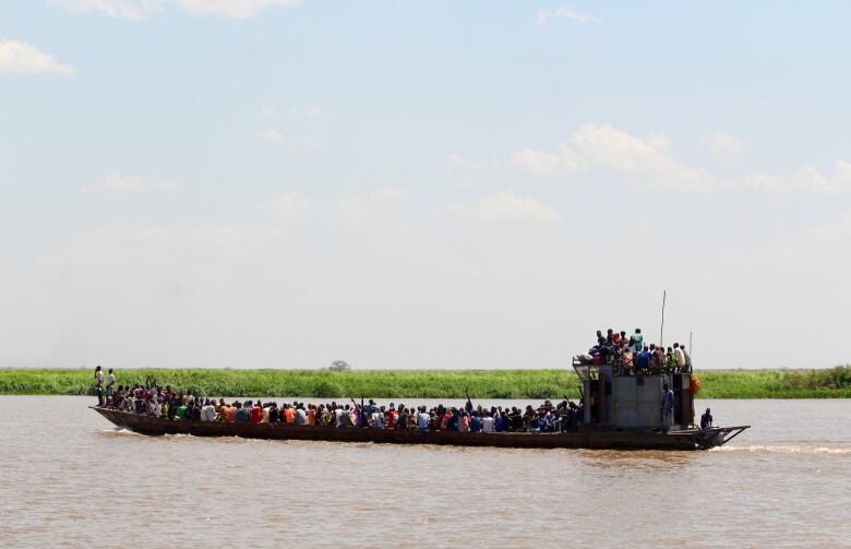 A boat full of people moves along a river. A grassy shore is seen on one side.