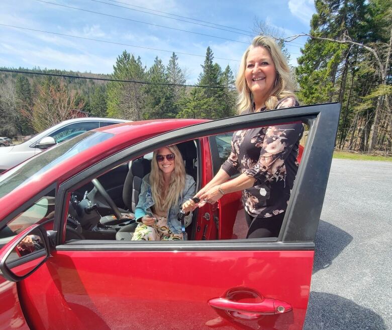 A young woman with long blonde hair sits in a red car, smiling and holding both hands in a 