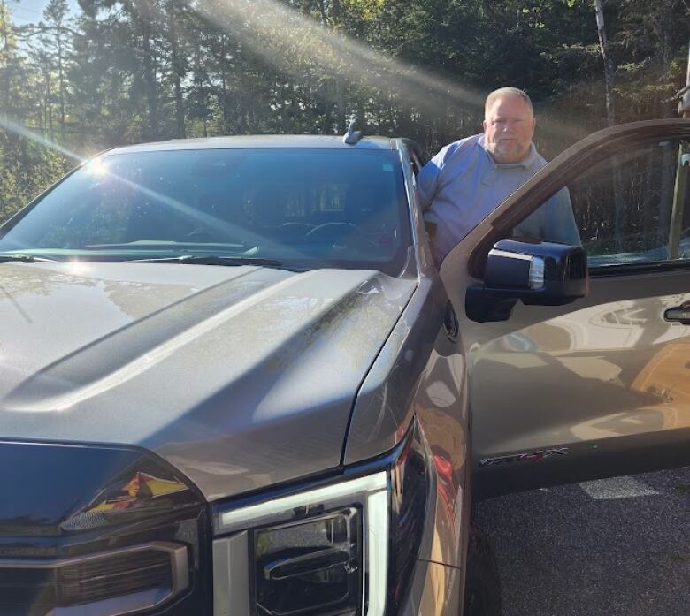 A man in a grey polo shirt peaks out from the open driver side door of a silver-grey pickup truck.