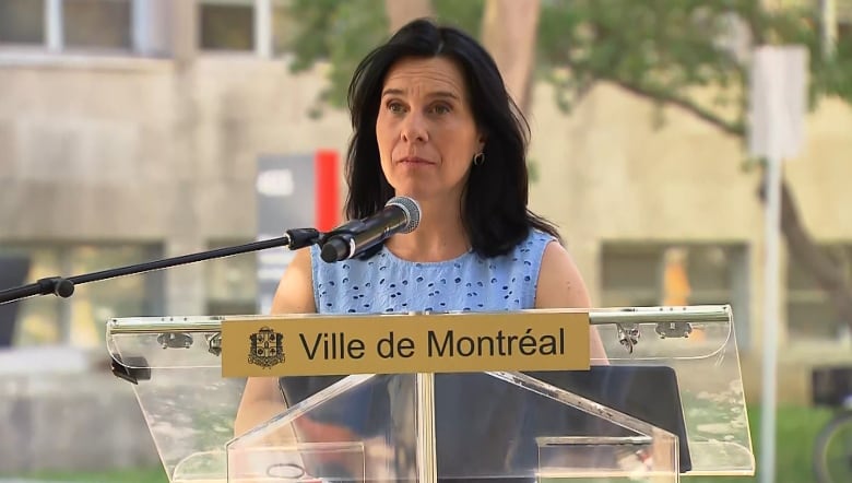 Montreal Mayor Valrie Plante stands at a clear city podium at an outdoor news conference on a hot sunny day, wearing a light blue top.