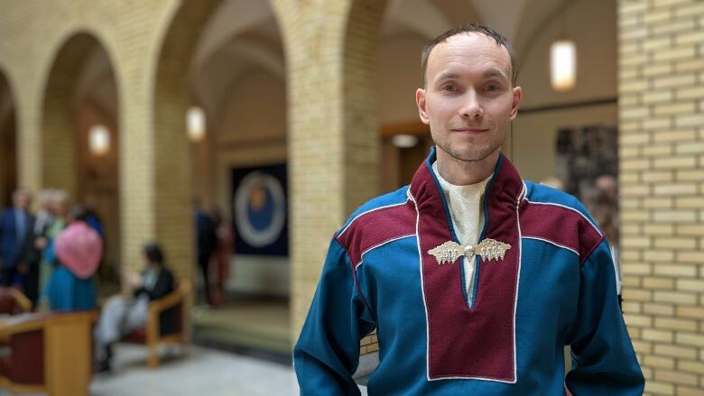 A man wearing a blue and maroon pullover, with an ornate clasp at the neck, poses for a portrait inside a brick building.