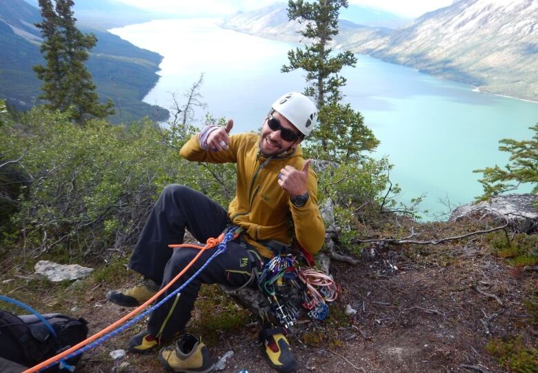 A man in rock-climbing gear gives a thumbs-up on a ledge overlooking a lake in the mountains. 