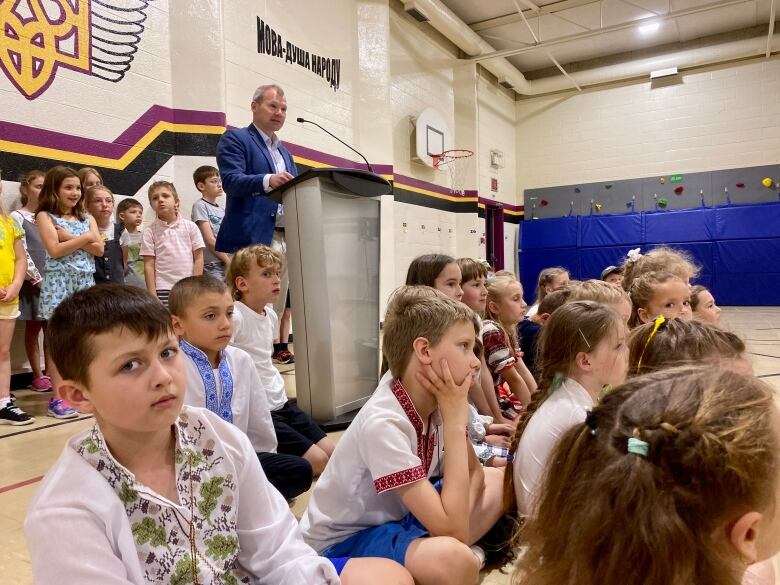 Children sitting in a gym as a man in a blue suit speaks. 