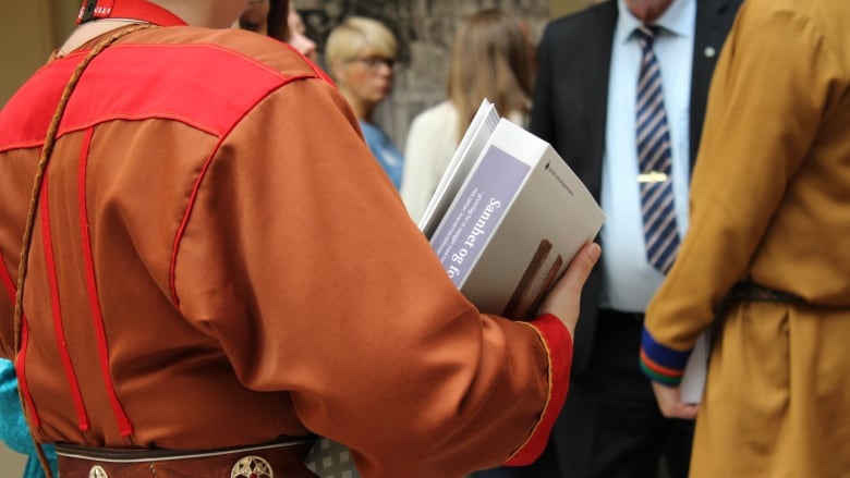 A person wearing a brown and red jacket and belt stands with their back to the camera, carrying a thick booklet in the crook of their right arm.