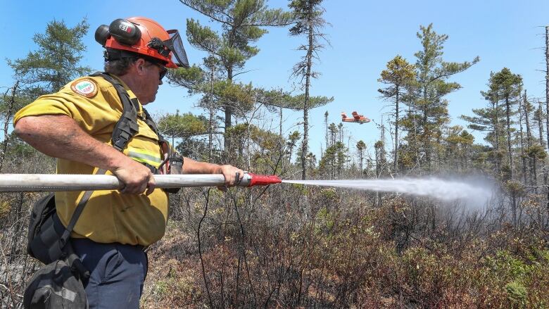 A man shoots water from a fire hose at a fire while a water bomber aircraft flies in the background.