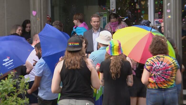 A man in a grey school talks to a crown of pride supporters. 