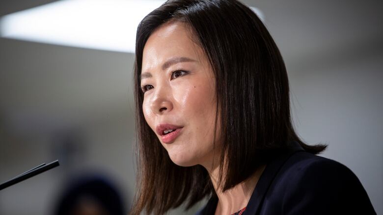 An Asian woman is shown from the shoulders up in front of a microphone. She has a black bob that hangs just at her shoulders and is wearing a navy blazer and a red shirt underneath. 