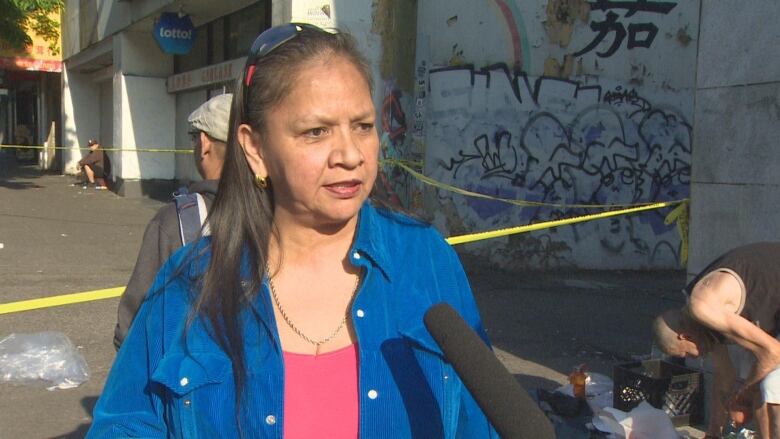 A woman with long brown hair and a blue shirt stands speaking outside a business storefront with graffiti.