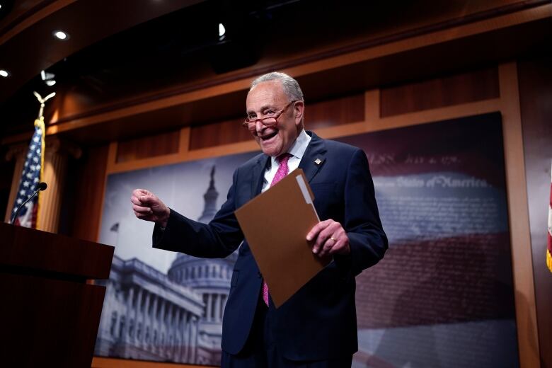 A man in a business suit and glasses smiles as he holds a file folder and stands near a podium.