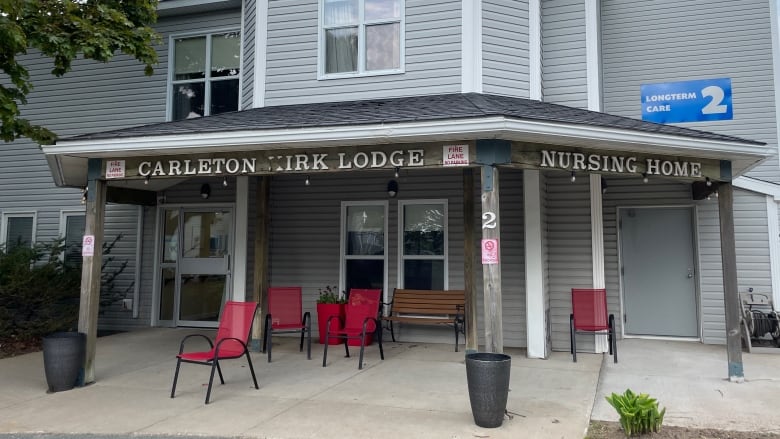 The exterior of the front of a building with grey siding, and a seating area, with a wooden bench and four red stackable patio chairs.