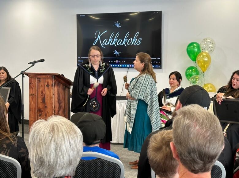 Two women stand and sing at the front of a room full of people.