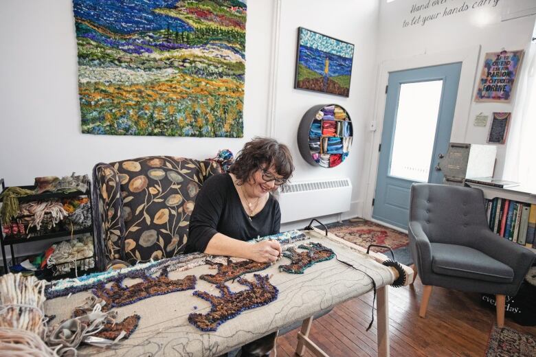 Deanne Fitzpatrick works on a rug covered in brown lambs in her studio. The cozy studio has art on the walls. Fitzpatrick sits in a large black chair with a floral pattern.