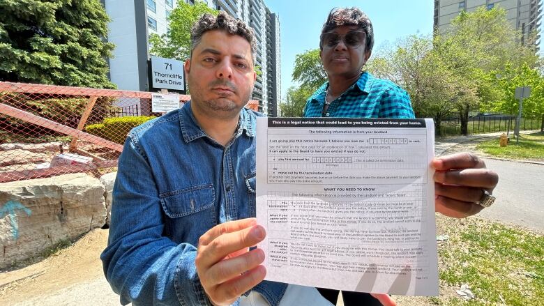 A man and a woman hold up a notice of eviction in front of an apartment building. 