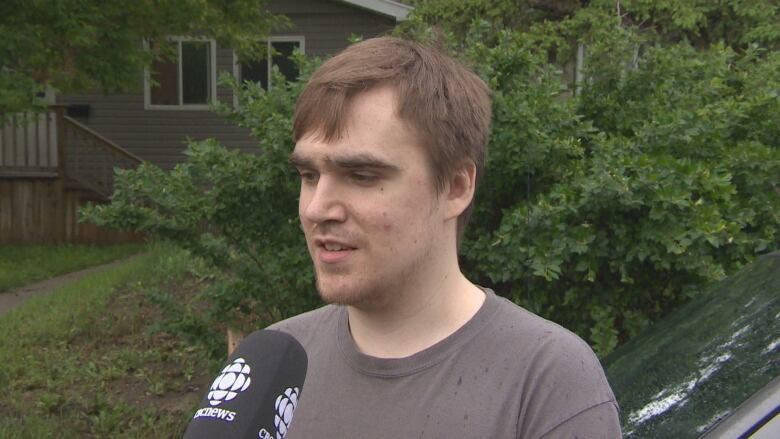 A person is standing beside a car and there is green shrubbery behind him and there is a CBC microphone just below his mouth.