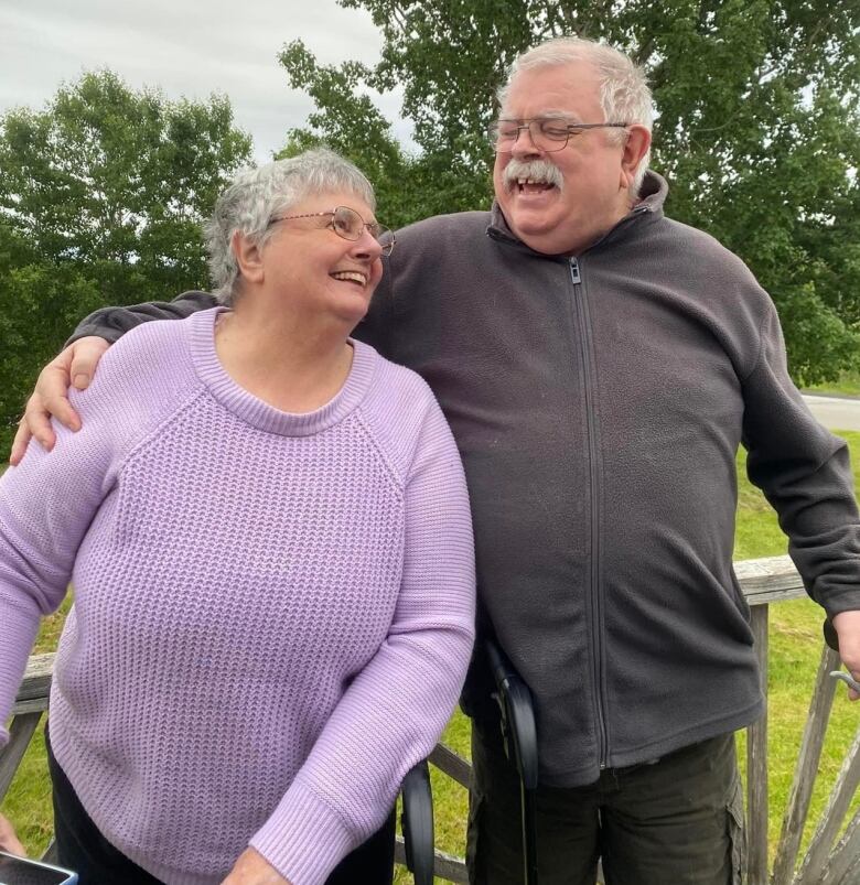 A man with grey hair, moustache and glasses has his right around around the shoulders of a woman with grey hair and glasses, as they look at each other, smiling.