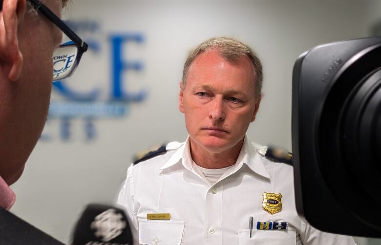 A man in a white police shirt stands in front of a reporter and a camera. 