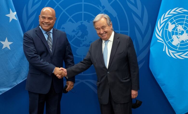 Two men shake hands in front of blue UN flag