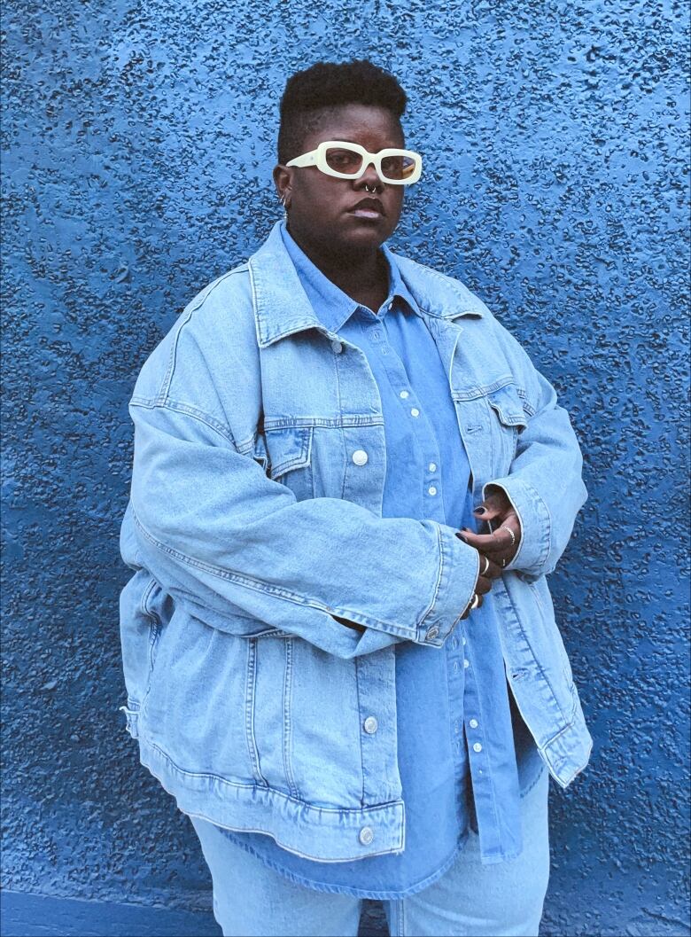 A person is pictured wearing an all-denim stylish blue outfit with white sunglasses, standing in front of a blue wall. 