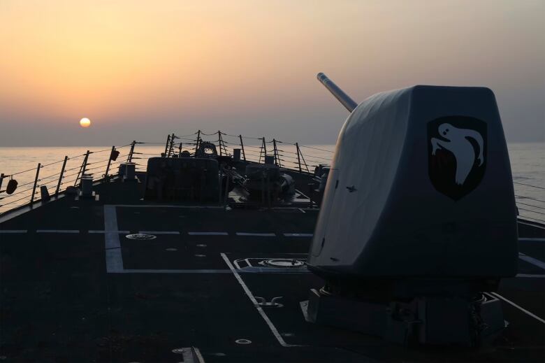 A large gun is visible on the front of a large ship. The sun hangs low in the sky.