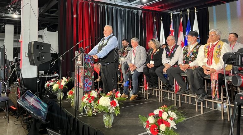 A man is pictured speaking at a podium. Flags and people seated in 2 rows are behind him on stage.