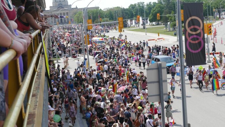 A crowd of people, many holding rainbow flags, is walking down a street. There are other people watching from the upper floors of a nearby parkade.